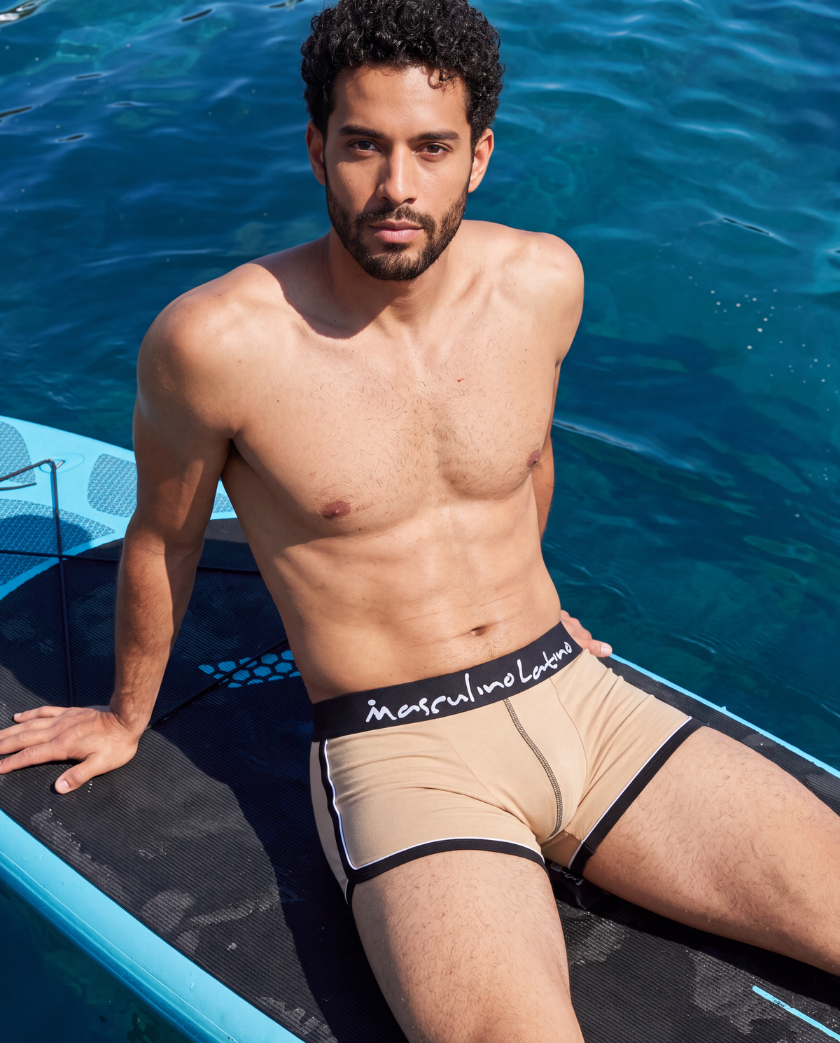 A man sitting on a blue paddleboard in front of a swimming pool, wearing beige briefs with black trim and a black waistband featuring the white script logo 'intrful me label'.